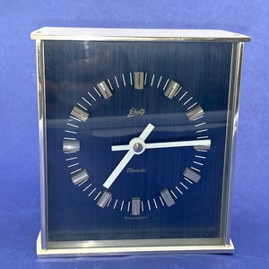 May include: A vintage Schatz Elacda clock with a rectangular silver frame and a dark blue face. The clock face features white hands and markers, with the brand name "Schatz" and "Elacda" displayed. The clock is a classic design.