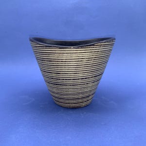 May include: A decorative vase with a unique shape and design. The vase has a dark brown rim and is covered in horizontal stripes of alternating light beige and dark brown. The vase is set against a blue background.