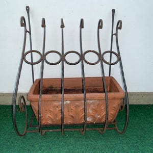 Può includere: Una fioriera rettangolare in terracotta con un motivo a foglie in rilievo, inserita in una struttura in ferro battuto marrone scuro. Il telaio presenta barre metalliche verticali ritorte, accenti circolari e decorazioni a volute.