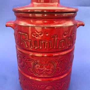 Könnte beinhalten: Ein roter Keramik-Rumtopf mit Deckel und Griffen. Der Topf hat geprägte Schriftzüge und Fruchtdesigns. Der Topf ist zylindrisch und hat eine glänzende Oberfläche. Der Hintergrund ist einfarbig blau.