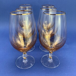 May include: Six clear wine glasses with gold rims and gold glitter accents. The glasses have a clear stem with a gold accent and a round base. The glasses are arranged on a blue background.