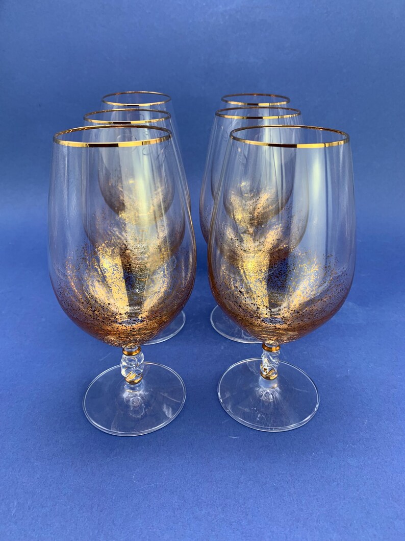 May include: Six clear wine glasses with gold rims and gold glitter accents. The glasses have a clear stem with a gold accent and a round base. The glasses are arranged on a blue background.