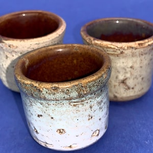 Può includere: Tre piccoli vasi in ceramica fatti a mano con un aspetto rustico e macchiettato. I vasi hanno una forma cilindrica con un bordo leggermente svasato. Gli interni sono di un marrone intenso, mentre gli esterni sono un mix di bianco, marrone e verde-blu.