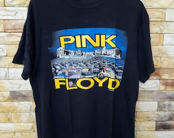 May include: Black t-shirt featuring the words "PINK FLOYD" in yellow, with a blue background and a graphic of a crowd of people. The shirt is hanging on a wooden hanger against a brick wall.