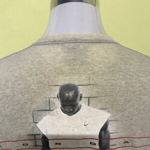 Op de afbeelding: Grijs T-shirt met een grafische print van een basketballer voor een bakstenen muur. De grafiek heeft het woord "JORDAN" in rood onderaan gedrukt.