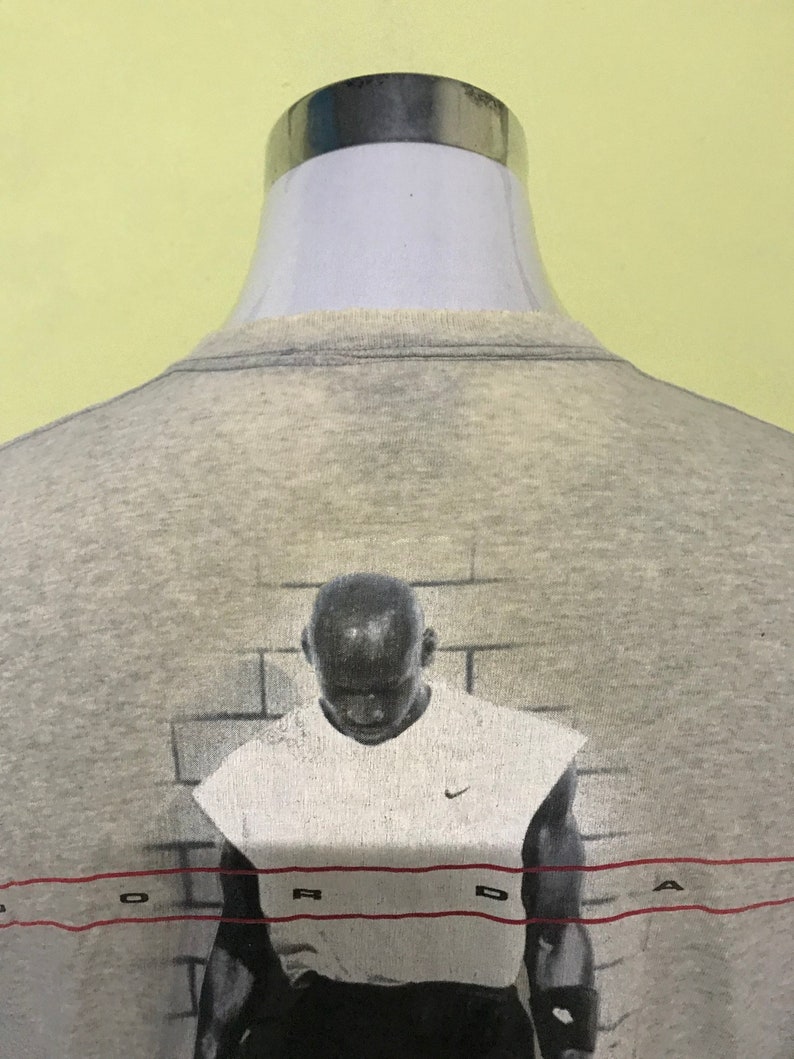 Op de afbeelding: Grijs T-shirt met een grafische print van een basketballer voor een bakstenen muur. De grafiek heeft het woord "JORDAN" in rood onderaan gedrukt.