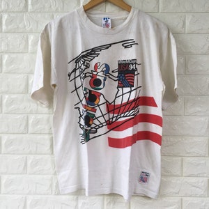 May include: White t-shirt with a graphic of a world map and the text "World Cup USA '94". The graphic is in black, red, blue, green, and yellow. The shirt has three red stripes on the bottom right side.