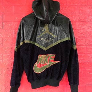 Könnte beinhalten: Schwarze Kapuzenjacke mit einem Nike-Logo und dem Wort "Flight" unter einem Jumpman-Logo. Die Jacke hat eine schwarze Lederkapuze und Schultern mit einem grün-schwarzen Design auf Brust und Ärmeln.