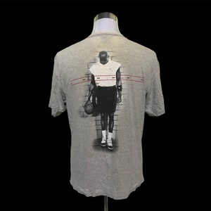 Op de afbeelding: Grijs T-shirt met een zwart-witte afbeelding van een basketballer die een basketbal vasthoudt. De tekst "JORDAN" is in rood op de achterkant van het shirt gedrukt.