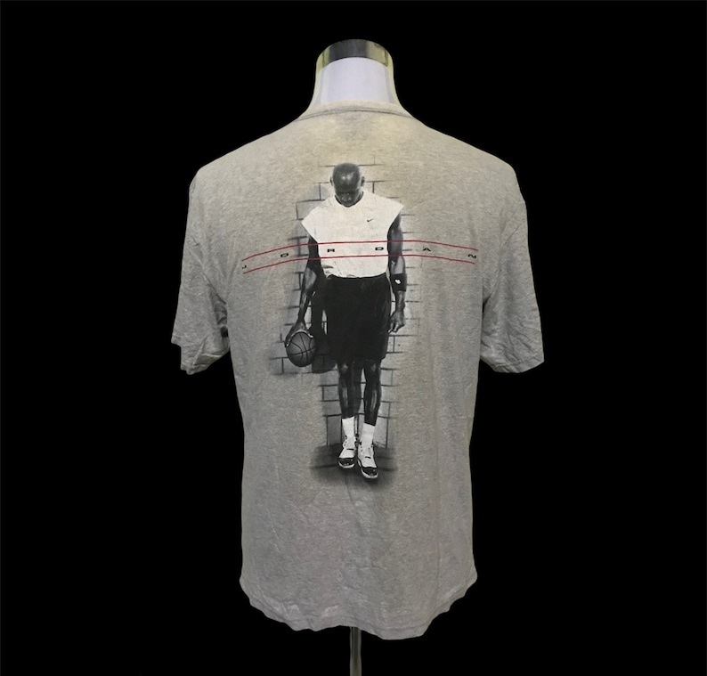 Op de afbeelding: Grijs T-shirt met een zwart-witte afbeelding van een basketballer die een basketbal vasthoudt. De tekst "JORDAN" is in rood op de achterkant van het shirt gedrukt.