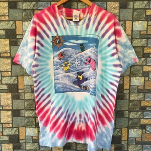 May include: Tie-dye t-shirt with a colorful graphic of the Grateful Dead bears snowboarding in a snowy mountain scene. The shirt has a blue, pink, and white swirl pattern.