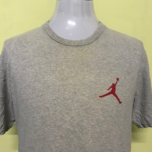 Op de afbeelding: Een grijs T-shirt met een rood silhouet van een basketballer die in de lucht springt. De speler houdt een basketbal in zijn rechterhand.