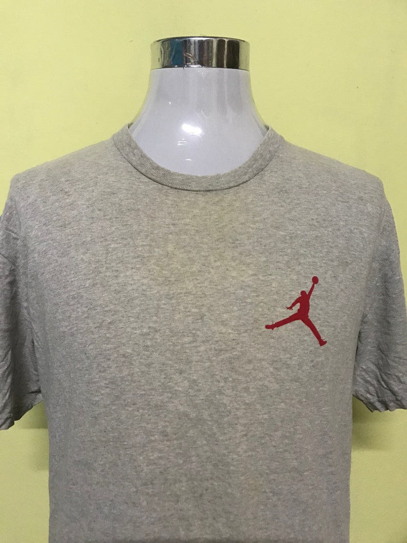 Op de afbeelding: Een grijs T-shirt met een rood silhouet van een basketballer die in de lucht springt. De speler houdt een basketbal in zijn rechterhand.