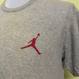 Op de afbeelding: Een grijs T-shirt met korte mouwen met een rood silhouet van een basketballer die in de lucht springt.