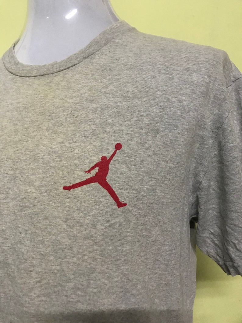 Op de afbeelding: Een grijs T-shirt met korte mouwen met een rood silhouet van een basketballer die in de lucht springt.