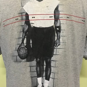 Op de afbeelding: Een grijs T-shirt met een zwart-witte afbeelding van een basketballer die een basketbal vasthoudt. De speler draagt een wit shirt zonder mouwen, een zwarte broek en witte sneakers. De tekst "JORDAN" is in rood bovenaan het shirt gedrukt.