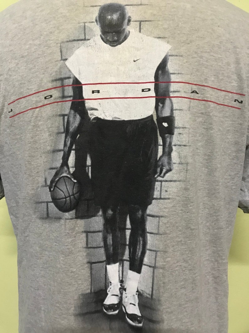 Op de afbeelding: Een grijs T-shirt met een zwart-witte afbeelding van een basketballer die een basketbal vasthoudt. De speler draagt een wit shirt zonder mouwen, een zwarte broek en witte sneakers. De tekst "JORDAN" is in rood bovenaan het shirt gedrukt.