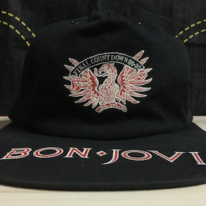 Peut inclure: Casquette de baseball noire avec un aigle brodé en rouge et blanc et le texte "Final Count Down 90-91". Le devant de la casquette porte le texte "Bon Jovi" en rouge et blanc.