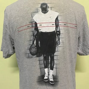 Op de afbeelding: Grijs T-shirt met een zwart-witte afbeelding van een basketballer die een basketbal vasthoudt. De speler draagt een wit tanktop en een zwarte short. De tekst "JORDAN" is in rood boven de afbeelding gedrukt.