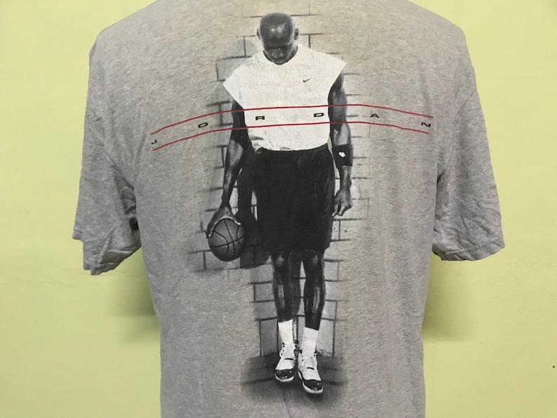Op de afbeelding: Grijs T-shirt met een zwart-witte afbeelding van een basketballer die een basketbal vasthoudt. De speler draagt een wit tanktop en een zwarte short. De tekst "JORDAN" is in rood boven de afbeelding gedrukt.