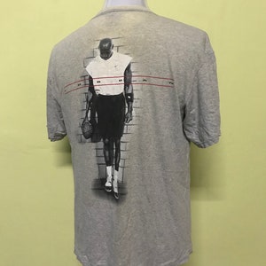 Op de afbeelding: Een grijs T-shirt met een zwart-witte afbeelding van een basketballer op de achterkant. De speler draagt een wit tanktop en een zwarte broek. De tekst "JORDAN" is in rood gedrukt op de achterkant van het shirt.