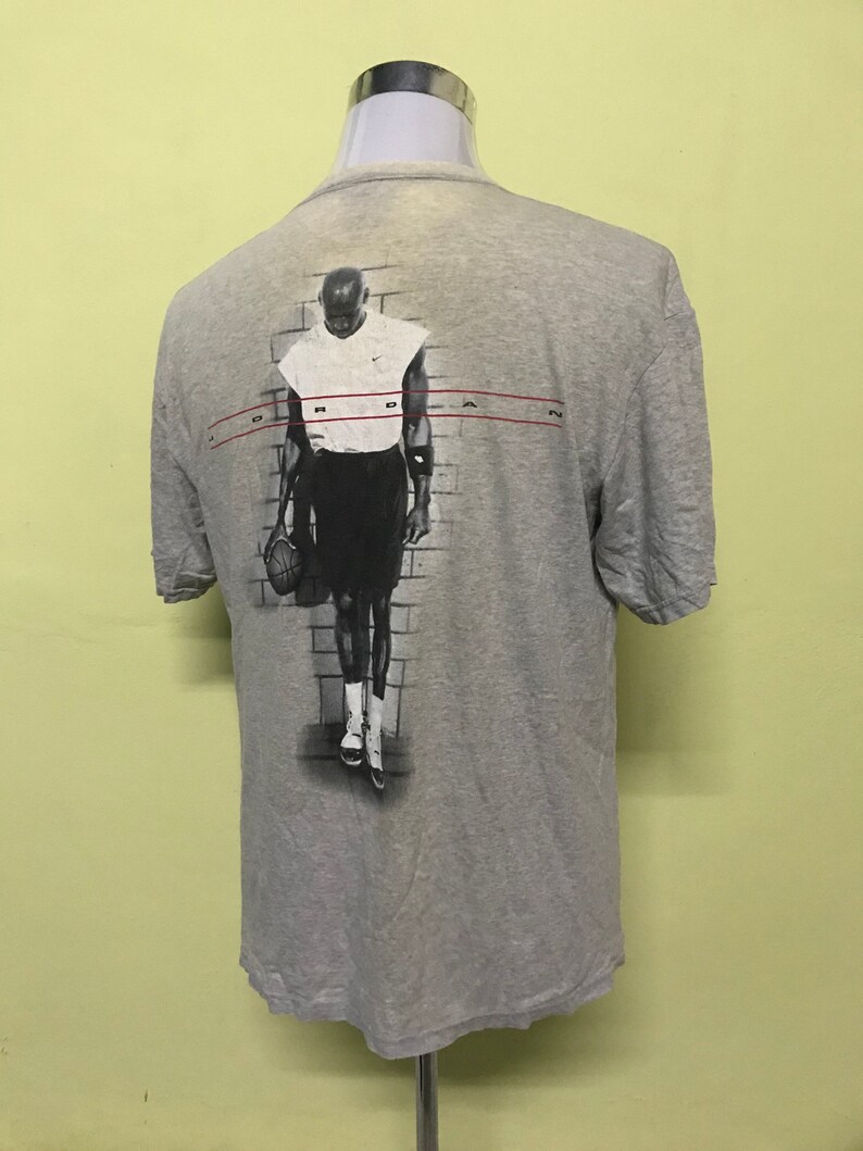 Op de afbeelding: Een grijs T-shirt met een zwart-witte afbeelding van een basketballer op de achterkant. De speler draagt een wit tanktop en een zwarte broek. De tekst "JORDAN" is in rood gedrukt op de achterkant van het shirt.