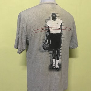 Op de afbeelding: Grijs T-shirt met een grafische print van een basketballer die een basketbal vasthoudt. De grafiek is in zwart-wit en heeft de tekst "JORDAN" bovenaan.