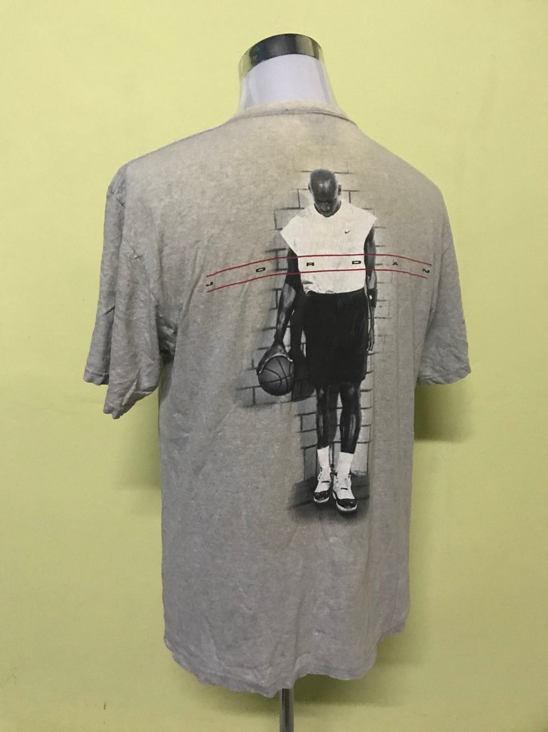 Op de afbeelding: Grijs T-shirt met een grafische print van een basketballer die een basketbal vasthoudt. De grafiek is in zwart-wit en heeft de tekst "JORDAN" bovenaan.