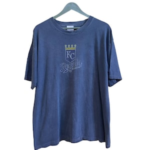 Può includere: Una maglietta vintage blu con una grafica sbiadita del logo dei Kansas City Royals. Il logo presenta una corona, le lettere "KC" e la parola "Royals" in un font corsivo.