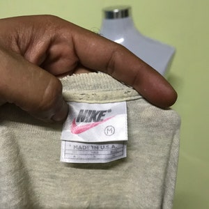 Op de afbeelding: Een wit Nike T-shirt met een rood Nike-logo en de tekst "MADE IN U.S.A." op een wit label. Het label heeft ook een maat "M" erop gedrukt.