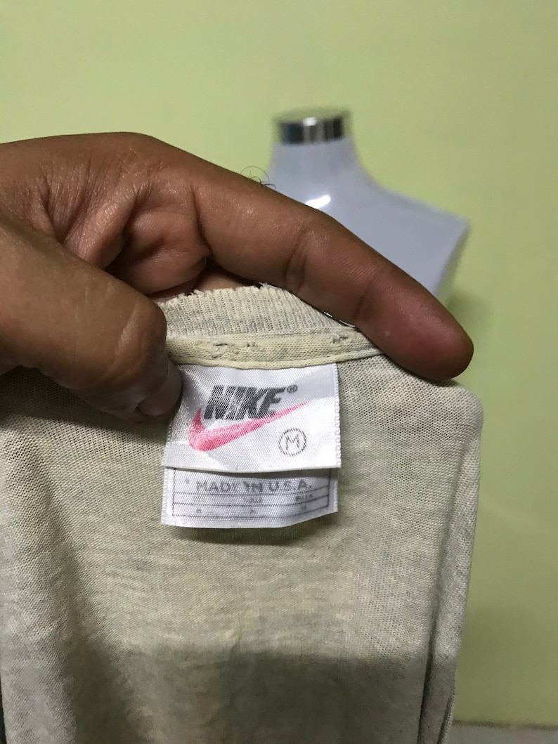 Op de afbeelding: Een wit Nike T-shirt met een rood Nike-logo en de tekst "MADE IN U.S.A." op een wit label. Het label heeft ook een maat "M" erop gedrukt.
