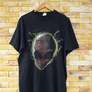 Puede incluir: Camiseta negra con un gráfico de extraterrestre verde y rojo. El extraterrestre está rodeado de rayos verdes.