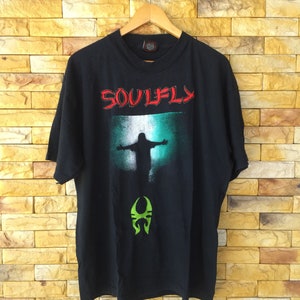 Könnte beinhalten: Schwarzes T-Shirt mit einem grünen und roten Aufdruck einer Band namens "Soulfly". Der Aufdruck zeigt eine Silhouette einer Person mit ausgestreckten Armen und einem grünen Symbol darunter.