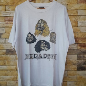 Może przedstawiać: Biały T-shirt z kreskówkową ilustracją zespołu Megadeth. Członkowie zespołu są przedstawieni z przesadzonymi cechami. Nazwa zespołu jest wydrukowana czarno-biało pod ilustracją.