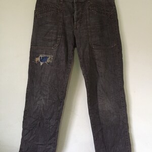 May include: Dark gray denim jeans with a distressed patch on the left leg. The jeans have large pockets on the front and back.