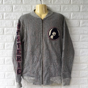 May include: A gray and black checkered zip-up jacket with a black and white patch on the left chest featuring a woman's face. The word "HYSTERIC" is embroidered in red and blue on the left sleeve.