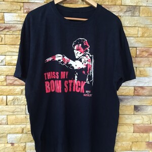 Può includere: Maglietta nera con una grafica rossa di un uomo che tiene una pistola e il testo "This my Bom Stick" e "Army of Darkness"