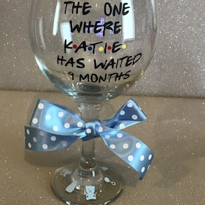 May include: A clear glass with a blue ribbon tied around the stem. The glass has the text "THE ONE WHERE KATIE HAS WAITED 9 MONTHS" written on it.