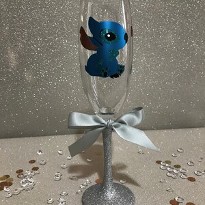 May include: A silver glitter champagne flute with a blue Stitch character decal. The flute has a silver glitter stem and a light blue bow.