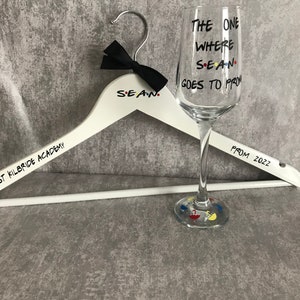 May include: A white wooden hanger with the text "Sean" and "St Kilbride Academy" printed on it. A black bow tie is attached to the hanger. A clear glass wine glass with the text "The One Where Sean Goes To Prom" and colourful dots is next to the hanger. The text "From 2022" is written on the hanger.