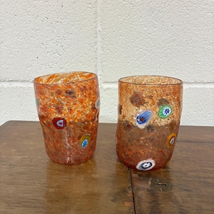 Peut inclure: Deux verres cylindriques présentant une base de couleur orange et ambre mouchetée avec des accents de verre millefiori multicolores incrustés. La surface de chaque récipient affiche un aspect texturé et tacheté avec des motifs circulaires.