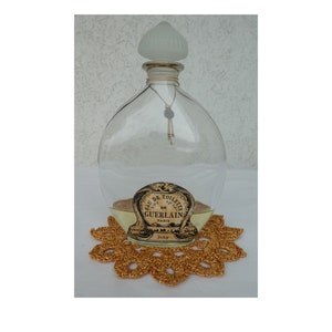 Könnte beinhalten: Eine klare Glasflasche mit einem mattierten Glasstopfen und einem goldenen Etikett mit der Aufschrift "Eau de Toilette de Guerlain Paris Jicky". Die Flasche steht auf einer goldenen Häkeldeckchen.