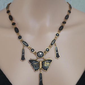 Op de afbeelding: Een gouden ketting met zwarte emaille hangers met Japanse sc&egrave;nes. Het middelpunt is een vlinderhanger met een gedetailleerde sc&egrave;ne van een Japanse tempel en een schip.