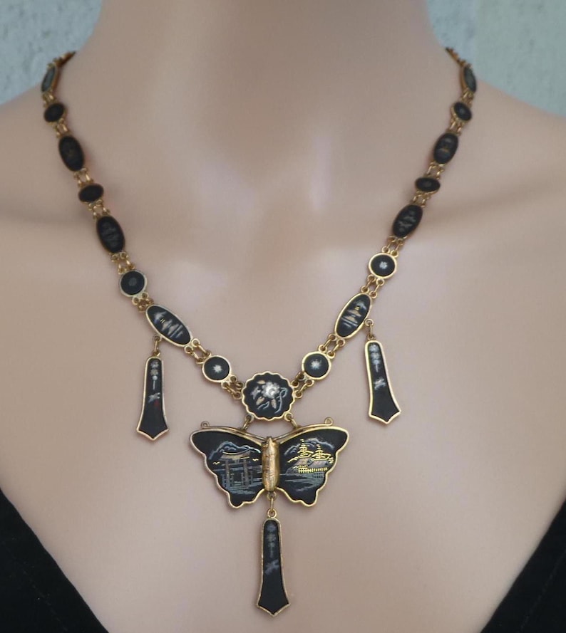 Op de afbeelding: Een gouden ketting met zwarte emaille hangers met Japanse sc&egrave;nes. Het middelpunt is een vlinderhanger met een gedetailleerde sc&egrave;ne van een Japanse tempel en een schip.