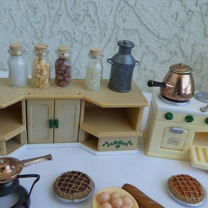 Puede incluir: Un juego de cocina en miniatura con un armario beige, una estufa blanca con una olla de cobre y una sartén de plata, y varios accesorios, incluyendo una lámpara, frascos y una jarra de leche.