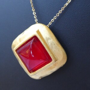 May include: A gold-toned necklace featuring a pendant with a textured, square-shaped frame. The pendant holds a vibrant, red, diamond-shaped stone. The necklace chain is delicate and gold-colored.