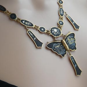 Op de afbeelding: Een gouden ketting met zwarte emaille hangers en een vlinderbedel. De vlinder heeft een zwart emaille lichaam met gouden accenten en witte details. De ketting is een statement piece met een uniek ontwerp.