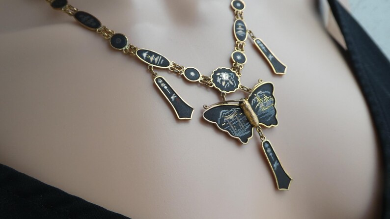 Op de afbeelding: Een gouden ketting met zwarte emaille hangers en een vlinderbedel. De vlinder heeft een zwart emaille lichaam met gouden accenten en witte details. De ketting is een statement piece met een uniek ontwerp.