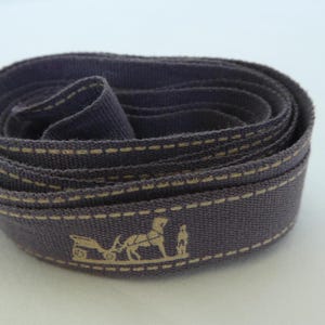 May include: A rolled-up dark blue fabric ribbon with a beige stitched edge. The ribbon has a repeating beige graphic of a horse-drawn carriage and a person. The ribbon is likely for decorative use, perhaps for gift wrapping.