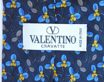 May include: A navy blue Valentino Cravatte tie with a repeating floral pattern. The design features blue flowers with yellow centers and gray leaves on thin stems. A white label with the Valentino logo and text is visible.